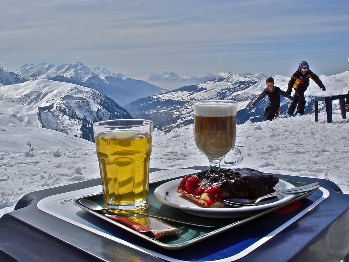 Kinder toben im Schnee vor Bergpanorama, im Vordergrund eine Pause auf der Skihütten-Terrasse